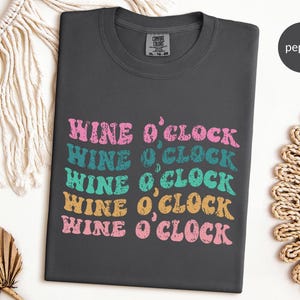 May include: A dark gray t-shirt with the text "Wine O'Clock" repeated five times in a retro font. The text is in pink, teal, and yellow.