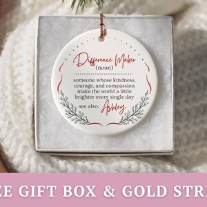May include: A white ceramic ornament in a gift box. The ornament has a gold string for hanging and features the words "Difference Maker" in red script, along with a definition and the name "Ashley". Decorative sprigs frame the text.