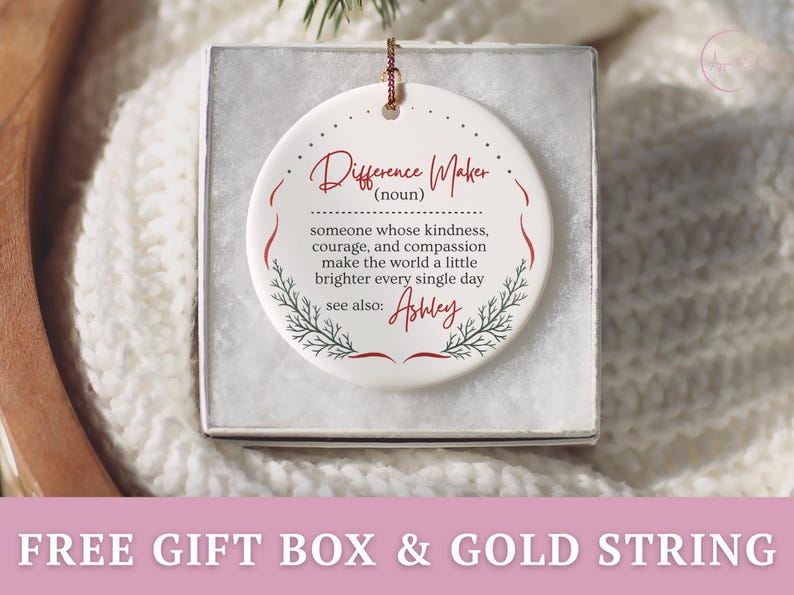 May include: A white ceramic ornament in a gift box. The ornament has a gold string for hanging and features the words "Difference Maker" in red script, along with a definition and the name "Ashley". Decorative sprigs frame the text.