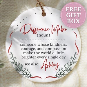 May include: White ceramic ornament with the text "Difference Maker" in red script. The ornament includes a definition and the name "Ashley". A pink circle with "FREE GIFT BOX" is in the upper right.