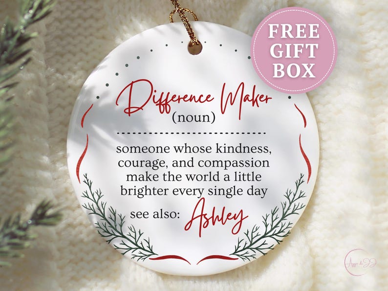 May include: White ceramic ornament with the text "Difference Maker" in red script. The ornament includes a definition and the name "Ashley". A pink circle with "FREE GIFT BOX" is in the upper right.