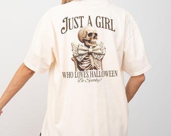 May include: Cream-colored t-shirt with a graphic of a skeleton wearing a bow tie. The text on the shirt reads "JUST A GIRL WHO LOVES HALLOWEEN Be Spooky!" The shirt has short sleeves and a relaxed fit.