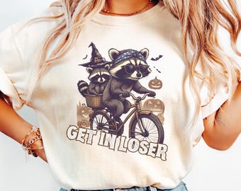 May include: Cream-colored t-shirt featuring a Halloween-themed graphic. The design includes two raccoons, one riding a bicycle and the other in a basket, with pumpkins and bats. The text "GET IN LOSER" is printed below the raccoons.