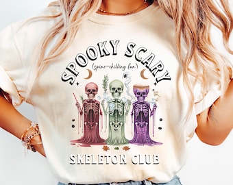 May include: Cream-colored t-shirt featuring a graphic of three skeletons in burgundy, green, and purple robes. The text "SPOOKY SCARY (spine-chilling fun)" and "SKELETON CLUB" are printed above and below the skeletons, respectively. The design includes moons, stars, and a spider.