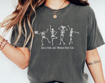 May include: Dark gray t-shirt featuring a white graphic of dancing skeletons. The text "Skeletons Just Wanna Have Fun" is printed below the skeletons. The shirt has a crew neck and short sleeves.