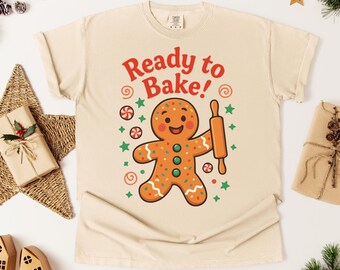 May include: Cream-colored t-shirt with a gingerbread man graphic holding a rolling pin. The text "Ready to Bake!" is in red above the gingerbread man. The gingerbread man has orange frosting and green and orange accents.