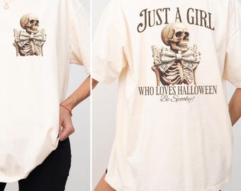 May include: Cream-colored t-shirt featuring a skeleton with a lace bow tie. The back of the shirt has the text "JUST A GIRL WHO LOVES HALLOWEEN Be Spooky!" in brown lettering. The shirt is a casual, oversized fit.