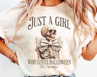 May include: Cream-colored t-shirt with the text "JUST A GIRL WHO LOVES HALLOWEEN Be Spooky!" featuring a detailed illustration of a skeleton with a bow tie. The design is centered on the shirt.