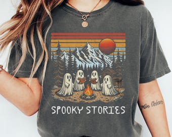 May include: Dark gray t-shirt with a graphic of ghosts around a campfire in a mountain scene. The design includes a sunset, trees, and the text "Spooky Stories" in white.