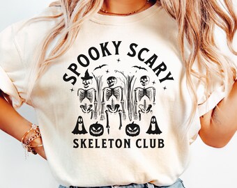 May include: Cream-colored t-shirt with the text "Spooky Scary Skeleton Club" in black. The design features three skeletons, bats, pumpkins, and ghosts, perfect for Halloween.