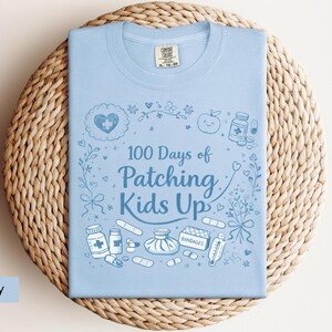 May include: Light blue t-shirt with the text "100 Days of Patching Kids Up" and medical-themed illustrations. The design includes a heart, apple, medicine bottles, bandages, and a thermometer. The shirt is folded on a woven, circular surface.