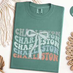 May include: A light green t-shirt with a retro-style graphic of the word "Charleston" repeated in a rainbow of colors. A white bow is centered on the graphic.