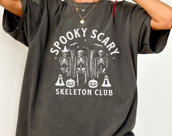 May include: Dark gray t-shirt with the text "SPOOKY SCARY SKELETON CLUB" in white. The graphic features three skeletons, ghosts, and pumpkins. The shirt has short sleeves and a relaxed fit, perfect for Halloween.