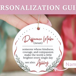 May include: A round, white ornament with the text "Difference Maker" in burgundy script. The ornament includes a definition and the name "Ashley" below. A gold string is attached for hanging. The background is blurred.