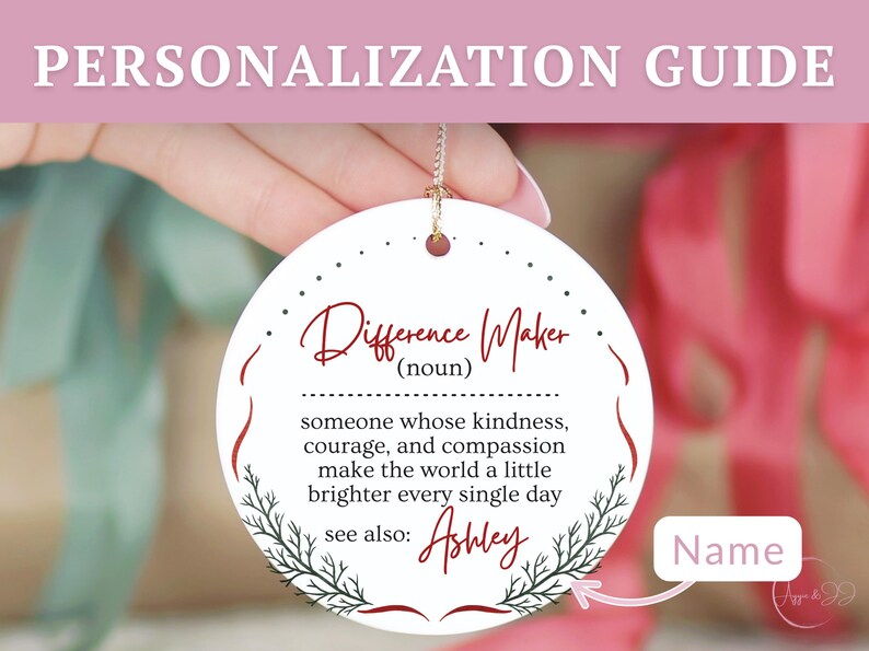 May include: A round, white ornament with the text "Difference Maker" in burgundy script. The ornament includes a definition and the name "Ashley" below. A gold string is attached for hanging. The background is blurred.