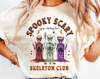 May include: Cream-colored sweatshirt with the words "SPOOKY SCARY" and "SKELETON CLUB" in brown and orange lettering. The graphic features three skeleton figures in maroon, green, and purple, with various accessories. A fun, Halloween-themed design.