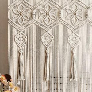 May include: Cream macrame wall hanging with geometric patterns and tassels. The design features diamond and floral motifs, with wooden beads at the center of the flowers. It is suspended from a wooden rod.