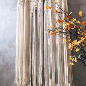 May include: Cream-coloured macrame curtains with detailed woven patterns and long, flowing fringe. The curtains hang from a wooden rod, creating a bohemian aesthetic. A branch with orange and yellow leaves is in the foreground.