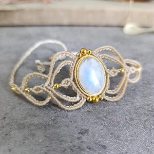 Könnte beinhalten: Handgefertigtes Makramee-Armband mit einem ovalen Mondstein in der Mitte. Das Armband ist aus beigem Kordel gefertigt und mit goldfarbenen Perlen verziert. Der Mondstein hat einen sanften, irisierenden Glanz, und das Design ist filigran.