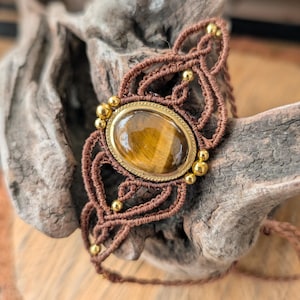Könnte beinhalten: Ein braunes Makramee-Armband mit einem großen, ovalen Tigeraugenstein, der in einen goldfarbenen Metallrahmen gefasst ist. Das Makramee-Design weist komplizierte Knoten auf und ist mit kleinen goldenen Perlen verziert. Das Armband wird auf Treibholz präsentiert.