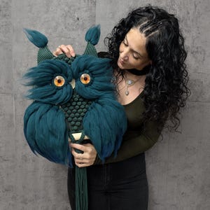 Puede incluir: Un colgante de pared con forma de búho, de color verde azulado y verde, con grandes ojos naranjas. El búho tiene plumas esponjosas de color verde azulado y un cuerpo de macramé. El búho tiene una larga borla verde.