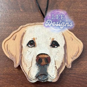 May include: A wooden dog ornament featuring a detailed carving of a golden retriever's face. The dog has brown eyes, a black nose, and a cream-colored coat. The ornament includes a black string for hanging and the text "LEE Designs".