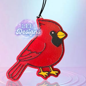 May include: A red cardinal bird-shaped ornament with black outlines and accents. The bird has a black face, yellow beak, and yellow feet. The ornament is hanging from a black string. The text "LEE Designs" is visible.