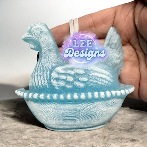May include: A light blue decorative hen ornament with the text "LEE Designs". The hen is sitting in a nest-like bowl. The ornament has a textured surface and a white ribbon for hanging.