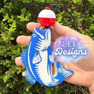 May include: A blue and white fish-shaped ornament featuring a red and white spherical fishing bobber attached to a black string. The ornament displays a textured surface with a graphic of a fish and the text LEE Designs in a purple circular logo.