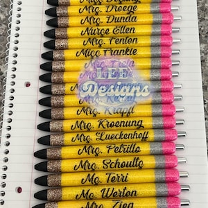 May include: A set of 18 yellow and pink glitter pens with black lettering. Each pen has a different name written on it, including "Miss Bohler", "Dr. Booker", "Miss Destiny", "Mrs. Droege", "Mig. Dunda", "Nurse Ellen", "Mrs. Fenton", "Miss Frankie", "Mrs. Trein", "Miss LEE", "Designs", "Mrs. Krapfl", "Mrs. Kroenung", "Mrs. Lueckenhoff", "Mrs. Petrillo", "Mrs. Schoulto", "Ms. Terri", "Mc. Werton", and "Mrs. Zieg".
