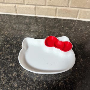 May include: A white, Hello Kitty-shaped dish with a red bow accent. The dish is made of a smooth material and is designed with a shallow, open area for holding small items. The bow is a bright red color.