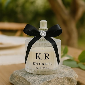 K&ouml;nnte beinhalten: Ein klares, geripptes Glas-Kerzenglas mit einer schwarzen Satin-Schleife und einem funkelnden Kristallakzent. Das Glas hat ein wei&szlig;es Etikett mit den Initialen "KR", den Namen "KYLE & RIEL" und dem Datum "10.05.2027". Die Kerze steht auf einem grauen Stein.