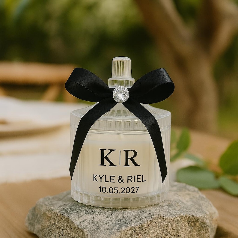 K&ouml;nnte beinhalten: Ein klares, geripptes Glas-Kerzenglas mit einer schwarzen Satin-Schleife und einem funkelnden Kristallakzent. Das Glas hat ein wei&szlig;es Etikett mit den Initialen "KR", den Namen "KYLE & RIEL" und dem Datum "10.05.2027". Die Kerze steht auf einem grauen Stein.