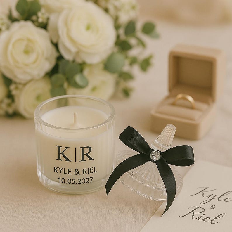 K&ouml;nnte beinhalten: Eine klare Glaskerze mit den Initialen "KR", den Namen "Kyle & Riel" und dem Datum "10.05.2027" aufgedruckt. Eine schwarze Schleife mit einem Kristallmittelst&uuml;ck verziert ein kleines Glasgef&auml;&szlig;. Ein goldener Ehering liegt in einer beigen Ringbox. Wei&szlig;e Blumen im Hintergrund.