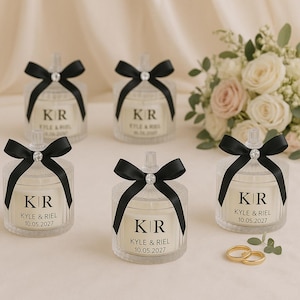 K&ouml;nnte beinhalten: Klare Glas-Kerzengl&auml;ser mit schwarzen Schleifen und Diamantakzenten. Jedes Glas hat die Initialen "KR" und die Namen "KYLE & RIEL" mit dem Datum "10.05.2027". Ein Blumenstrau&szlig; und zwei goldene Ringe sind im Hintergrund.