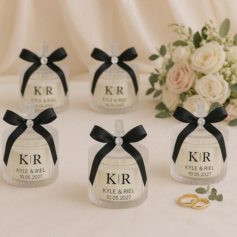 K&ouml;nnte beinhalten: Klare Glas-Kerzengl&auml;ser mit schwarzen Schleifen und Diamantakzenten. Jedes Glas hat die Initialen "KR" und die Namen "KYLE & RIEL" mit dem Datum "10.05.2027". Ein Blumenstrau&szlig; und zwei goldene Ringe sind im Hintergrund.