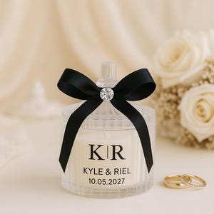 K&ouml;nnte beinhalten: Ein klares Glas-Kerzenglas mit einer schwarzen Schleife und einem funkelnden Strass-Akzent. Auf dem Glas sind die Initialen "KR", die Namen "KYLE & RIEL" und das Datum "10.05.2027" aufgedruckt. Zwei goldene Eheringe sind im Vordergrund.