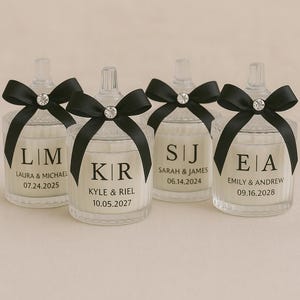 K&ouml;nnte beinhalten: Klare Glas-Kerzengl&auml;ser mit schwarzen Schleifen und Kristallakzenten. Jedes Glas hat personalisierten Text mit Initialen, Namen und Daten. Die Kerzen sind ein Hochzeits- oder Event-Gastgeschenk.