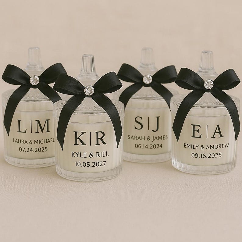 K&ouml;nnte beinhalten: Klare Glas-Kerzengl&auml;ser mit schwarzen Schleifen und Kristallakzenten. Jedes Glas hat personalisierten Text mit Initialen, Namen und Daten. Die Kerzen sind ein Hochzeits- oder Event-Gastgeschenk.