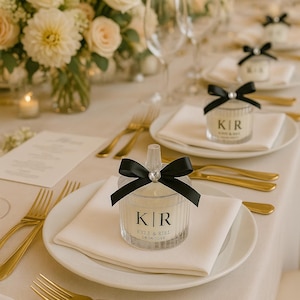 K&ouml;nnte beinhalten: Hochzeitstafel mit wei&szlig;en Tellern, Servietten und goldenem Besteck. Klare Kerzen-Gastgeschenke mit schwarzen Schleifen und den Initialen "K|R" sind auf jedem Teller platziert. Blumenschmuck und Kerzen erg&auml;nzen die elegante Dekoration.