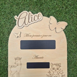 Peut inclure: Une plaque en bois en forme d'arche avec le prénom Alice en écriture cursive, deux papillons découpés et deux sections ardoise rectangulaires noires. Le texte gravé inclut Mon premier jour en et Année, avec des illustrations de faon et renard.