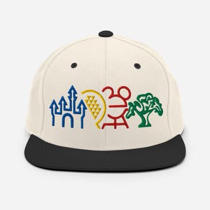 May include: A cream-colored snapback cap with a black brim and button top. The front features four embroidered icons in a row: a blue castle, a yellow crescent shape with a grid pattern, a red stylized mouse head, and a green tree silhouette.