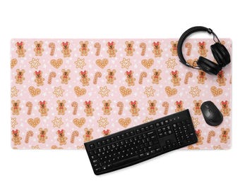 May include: A pink rectangular desk mat with a repeating pattern of gingerbread bears, hearts, snowflakes, and candy canes. A black keyboard, mouse, and headphones are on the mat. The mat is designed for a computer setup.