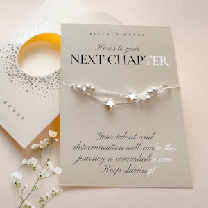 May include: A delicate silver-toned chain bracelet featuring small star charms and round beads, displayed on a beige card with the text "Here's to your NEXT CHAPTER" and an inspirational message about talent, determination, and keeping shining.