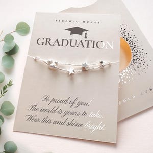 May include: A white card with the words "PICCOLO MUNDI" and "GRADUATION" printed on it. A silver charm bracelet with stars and beads is displayed on the card. The card also includes the text "So proud of you! The world is yours to take. Wear this and shine bright."