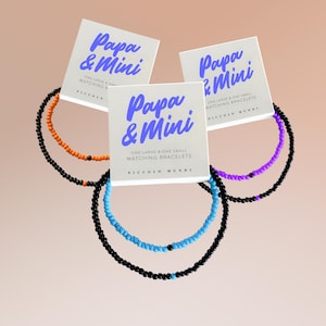 Può includere: Tre set di braccialetti abbinati, ciascuno con un braccialetto grande e uno piccolo, con la scritta "Papa & Mini". I braccialetti sono neri con perline arancioni, blu e viola.