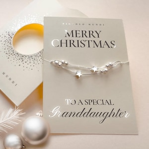 May include: A silver bracelet with star and bead accents is displayed on a Christmas card that reads "Merry Christmas To A Special Granddaughter." The card is light beige with silver lettering. A matching gift box and ornaments are in the background.
