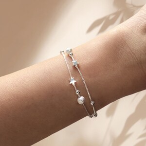 May include: Two silver bracelets with star and bead charms. The bracelets are layered on a person's wrist.