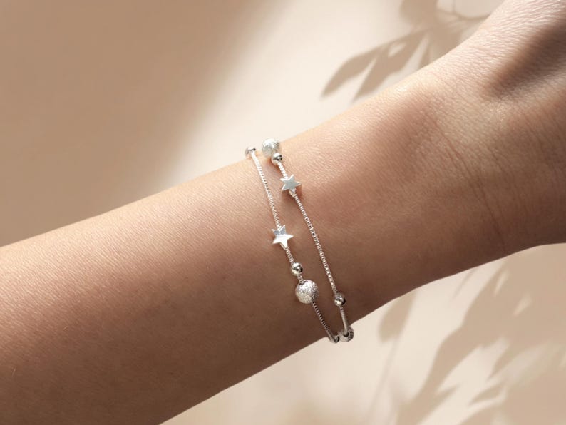 May include: Two silver bracelets with star and bead charms. The bracelets are layered on a person's wrist.