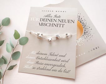 May include: A greeting card with a silver bracelet featuring star charms. The card has German text, including "Alles Gute für deinen neuen Abschnitt." A sprig of eucalyptus is in the background.